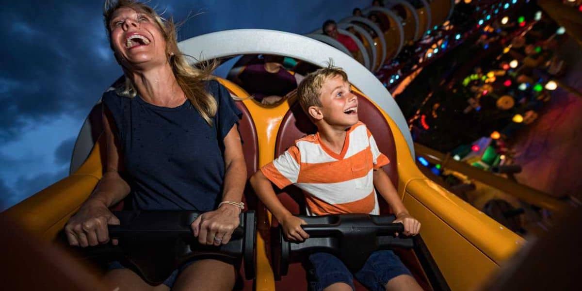 A woman and a young boy are seated on a roller coaster, both smiling and laughing with excitement. The ride is illuminated with colorful lights in the background, contrasting against a dark, cloudy sky.