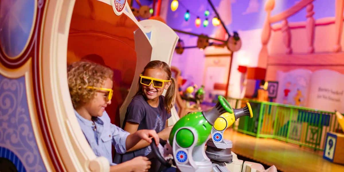 Two children wearing 3D glasses enjoy a ride at an amusement park. The background is colorful with playful decor and bright lights. Both are smiling and seated in a ride vehicle equipped with toy lasers.
