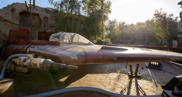 A Star Wars: Galaxy’s Edge red and white spaceship is on display outdoors, set among trees and rocky outcrops in the park.