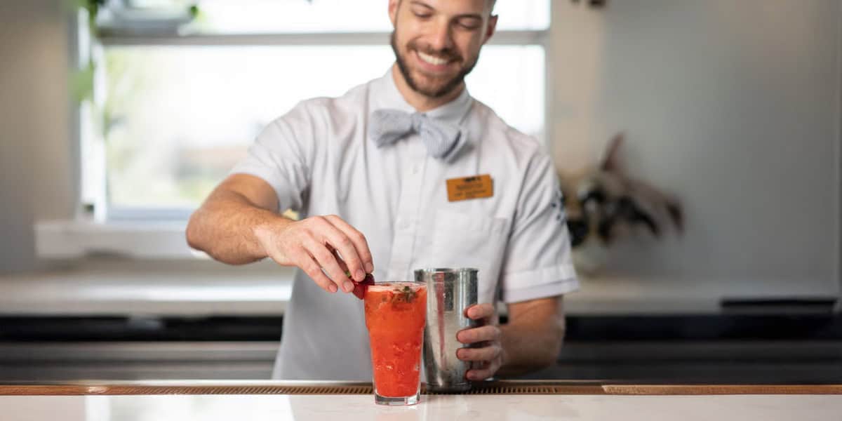 A smiling bartender at Paddlefish Disney Springs in a white shirt and bow tie garnishes a red cocktail with a strawberry at the bar counter, holding a metal shaker in his other hand.