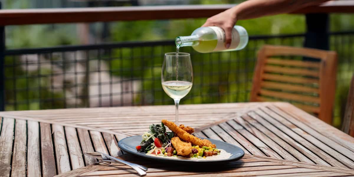 A hand pours a light-colored beverage into a wine glass on an outdoor wooden table at Paddlefish Disney Springs, with a plate of fried items and salad beside a fork and knife. Lush greenery and a railing complete the inviting scene.