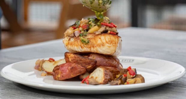 A grilled fish fillet topped with a vegetable medley, served over roasted red potatoes on a white plate at Paddlefish Disney Springs, with a savory sauce being drizzled on top.