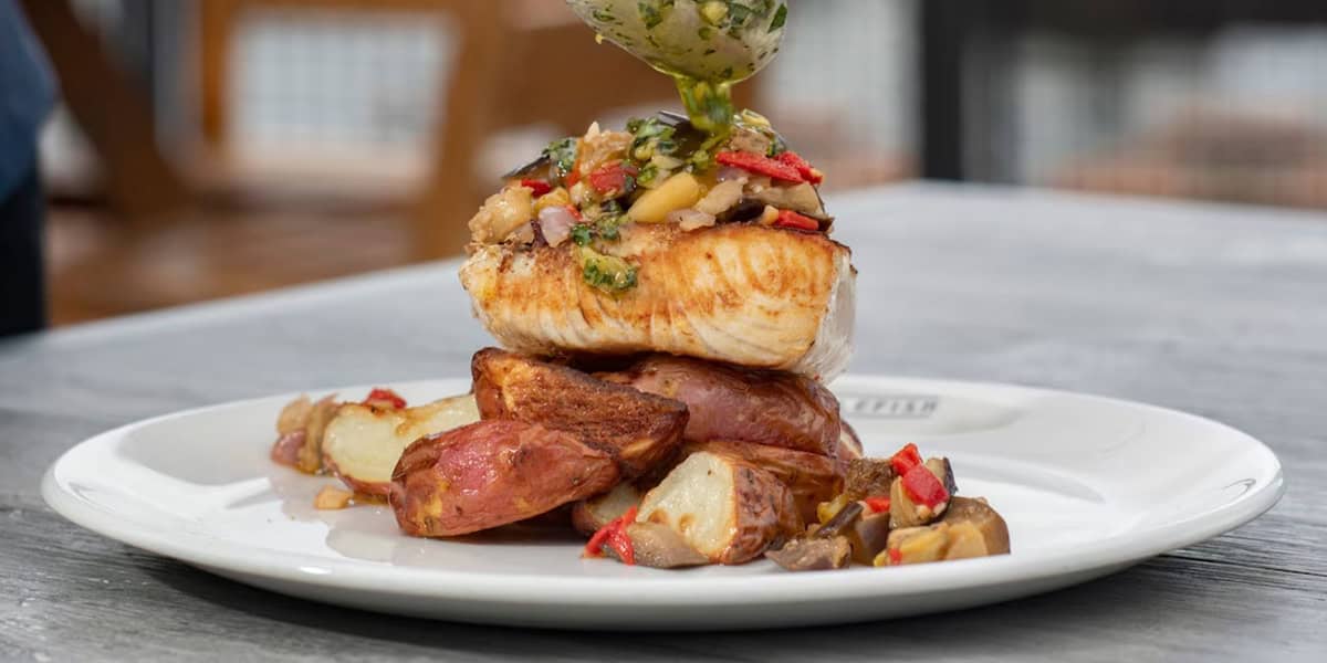 A grilled fish fillet topped with a vegetable medley, served over roasted red potatoes on a white plate at Paddlefish Disney Springs, with a savory sauce being drizzled on top.