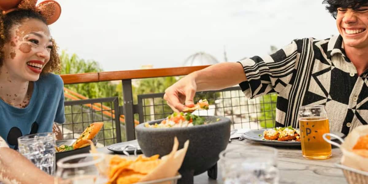 Two people smile and enjoy food and drinks at an outdoor Paddlefish Disney Springs restaurant table. One dips a chip into salsa while the other laughs, with plates and glasses visible on the table.
