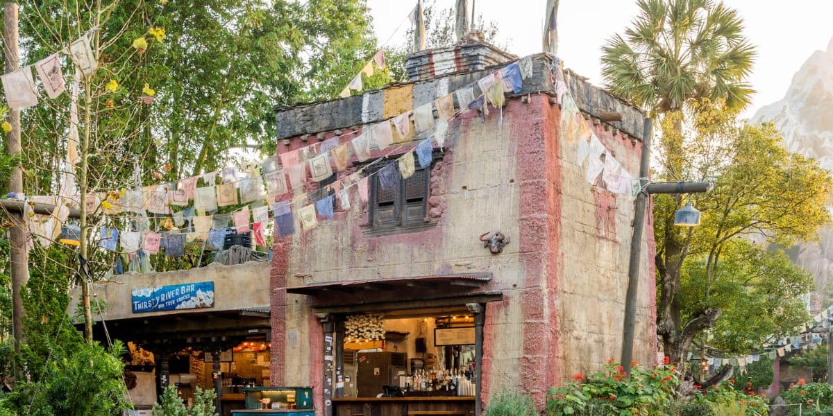 A rustic building with colorful prayer flags hanging above and around it. The structure has a small counter with drinks and snacks. Surrounded by lush greenery and trees, it exudes a tranquil, outdoor café vibe.