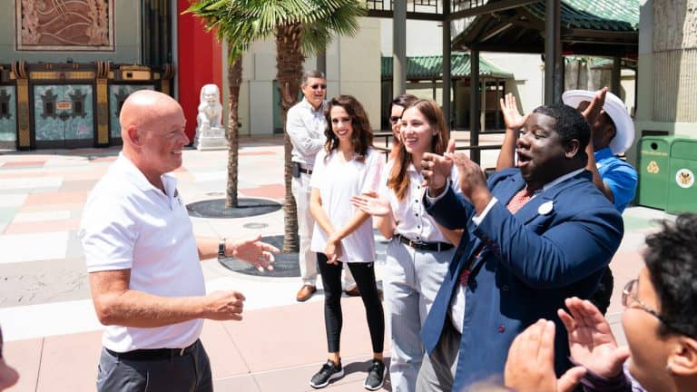 Guests cheer and applaud as a smiling bald man enjoys a sunny day surrounded by friends at Walt Disney World.