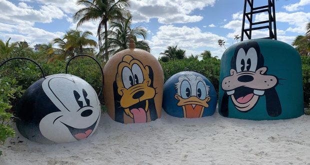 Whimsical Mickey, Pluto, Donald, and Goofy buoys decorate a Disney Cruise Line beach, nestled among swaying palm trees.