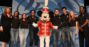 Mickey Mouse in a red band uniform joins a high school band and others on stage at Walt Disney World for a lively celebration.