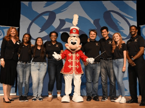 Mickey Mouse in a red band uniform joins a high school band and others on stage at Walt Disney World for a lively celebration.