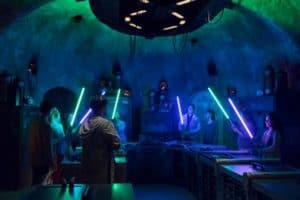 Guests wield glowing lightsabers in a futuristic Disney World setting illuminated by blue and green lights for an epic experience.