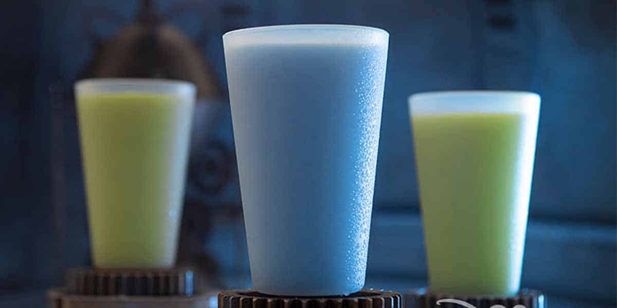 Three frosty drinks in opaque cups are displayed, reminiscent of Star Wars: Galaxy's Edge Food. The center cup contains a blue beverage, while the background cups feature a greenish-yellow drink. The softly blurred backdrop has a blue tone.