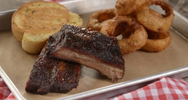 Barbecue ribs, onion rings, and garlic bread served at Regal Eagle Smokehouse in Epcot on a red checkered tray.