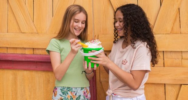 Two guests beaming as they share a famous Typhoon Lagoon ice cream sundae, topped with whipped cream, by a rustic boardwalk wall.