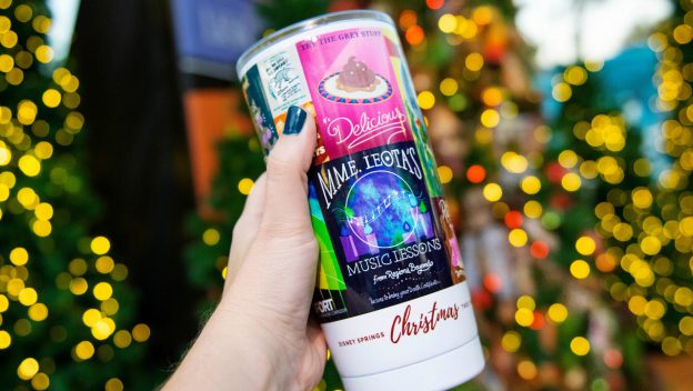 A festive holiday cup is held up with sparkling Christmas lights and elaborately decorated trees glowing in a theme park setting.