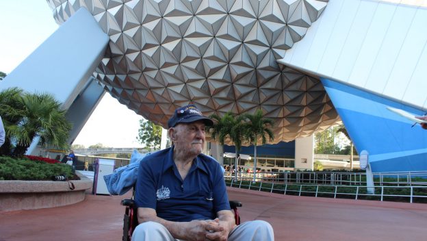 A WWII veteran in a wheelchair enjoys the view of Epcot’s Spaceship Earth, surrounded by Disney World’s lively atmosphere.