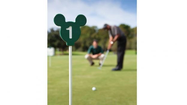 Mickey Mouse–themed green golf marker marked “1” on a Disney World course, with two golfers putting in the background.