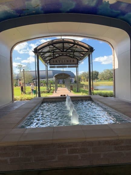 Fountain beneath an elegant arch at Disney’s Riviera Resort, inviting guests toward the Disney Skyliner on a bright, sunny day.