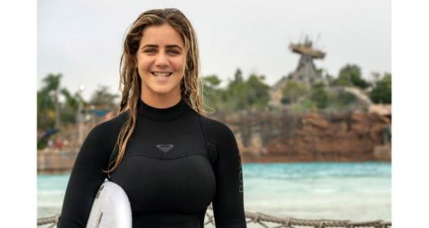 Olympic surfer Caroline Marks beams in a wetsuit, surfboard in hand, at Disney’s Typhoon Lagoon with its iconic wave pool backdrop.