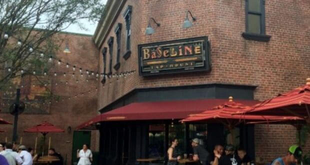 Outdoor view of Baseline Tap House, a brick building with a sign above the door. People are gathered under red umbrellas at tables. Trees and string lights enhance the casual atmosphere.