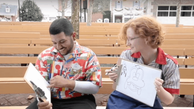 Fans challenge each other in a lively cartoon quick-draw contest, laughing and sketching on whiteboards amid theme park magic.