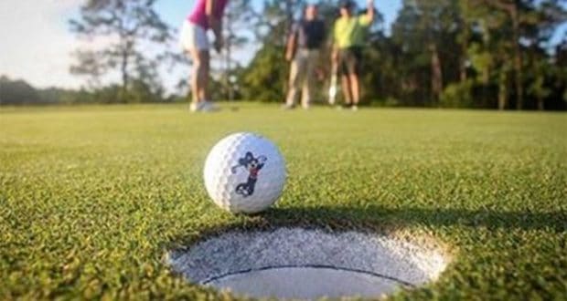 A golf ball teeters on the edge of a Disney Golf hole, with three excited guests enjoying the Player’s Club experience nearby.