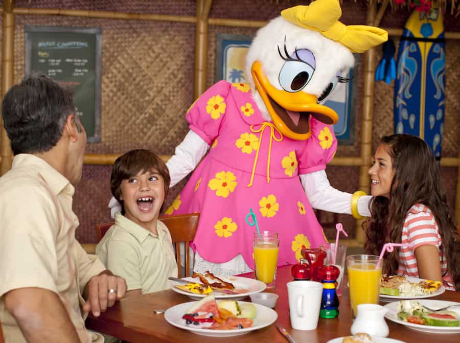 Kids and an adult share smiles with Daisy Duck in her pink dress at a vibrant Disney Parks restaurant, savoring magical moments.