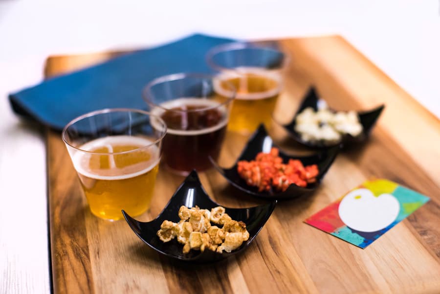 A flight of four beers with popcorn and snacks on a wooden board, set by a blue napkin and park-themed colorful card.
