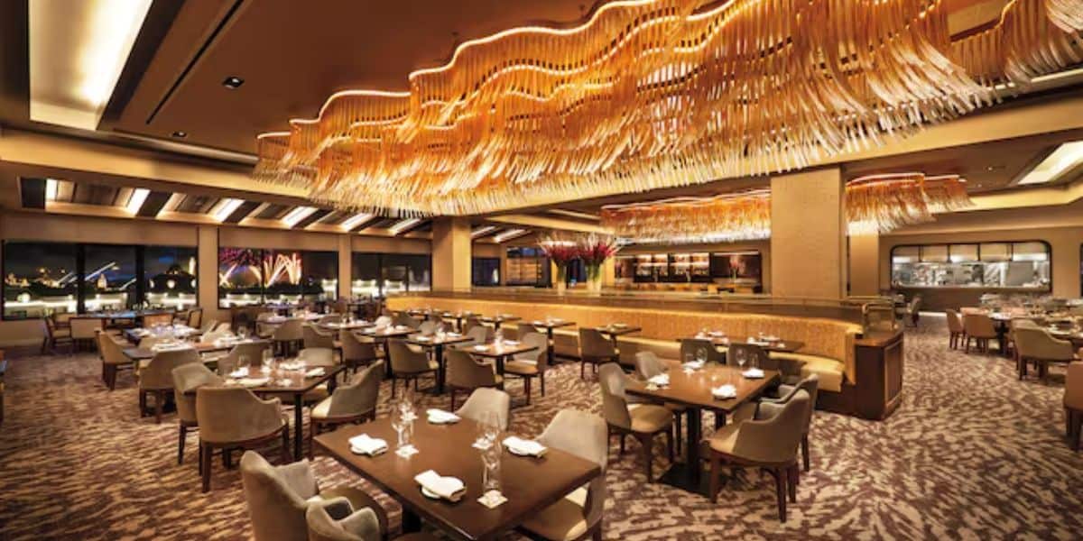 A luxurious restaurant interior featuring modern lighting with a wavy chandelier, neatly arranged tables with chairs, elegant decor, and large windows showcasing an evening cityscape view. Tables are set with glassware and napkins.