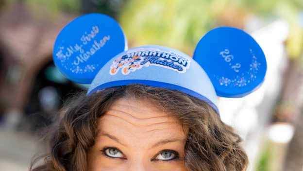 Disney fan beams in blue Mickey Mouse ears reading "Marathon Finisher" and "My First Marathon," celebrating runDisney victory.