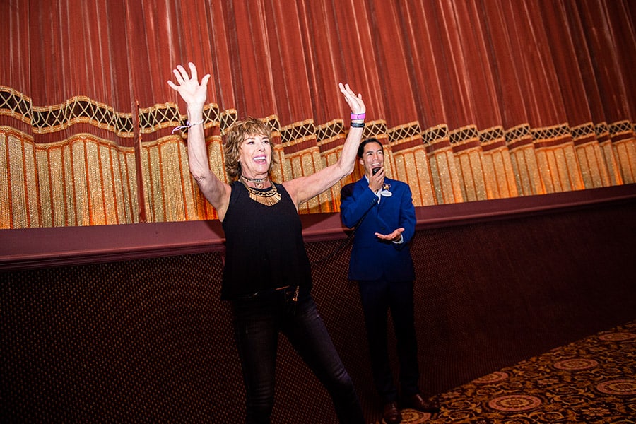 A guest raises her arms in excitement during a Beauty and the Beast Sing-Along as a Cast Member in a blue suit applauds behind her.