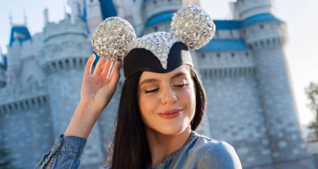 Disney fan beams in a sparkling Designer Collection Mickey Ear Hat, posing before the iconic castle at a Disney park.