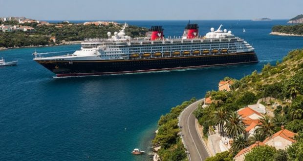 Disney Cruise Line ship glides by a scenic Greek town, red rooftops and green hills evoking the spirit of Disney adventures.