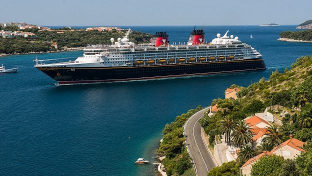 Disney Cruise Line ship glides by a scenic Greek town, red rooftops and green hills evoking the spirit of Disney adventures.