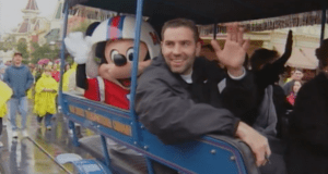 Kurt Warner and Mickey Mouse wave from a blue car during a Disney World parade, cheered on by raincoat-clad park guests.