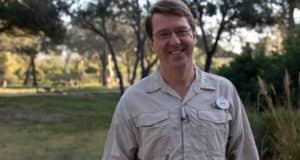 Dr. Mark Penning, wildlife expert from Disney’s Animal Kingdom, smiles outdoors in a sunny wooded area wearing his khaki shirt.