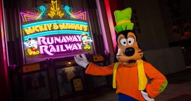 Goofy celebrates the dedication of Mickey & Minnie’s Runaway Railway, striking a pose by its glowing neon sign at night.