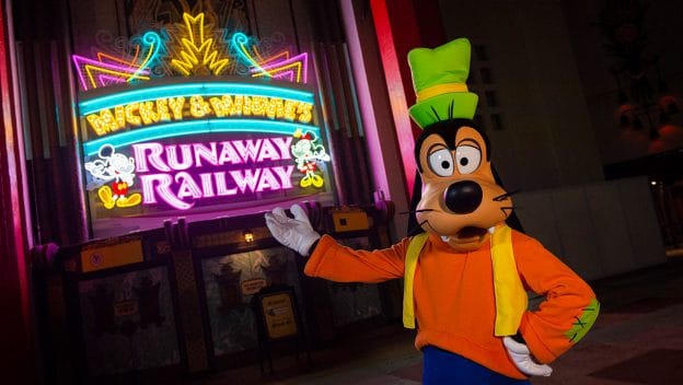 Goofy celebrates the dedication of Mickey & Minnie’s Runaway Railway, striking a pose by its glowing neon sign at night.