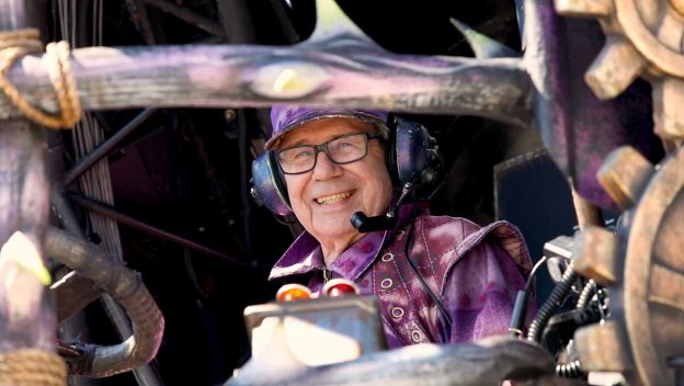 Festival of Fantasy Parade performer in vibrant purple costume and headset operates whimsical gears, bringing parade magic to life.