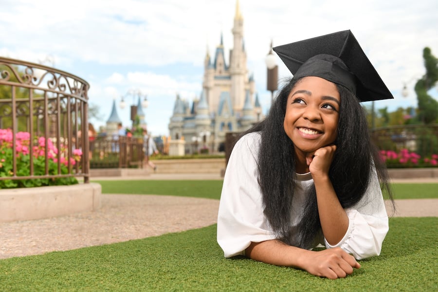 Graduate in cap relaxes on the grass near Cinderella Castle, celebrating Magic Kingdom graduation under sunny skies.