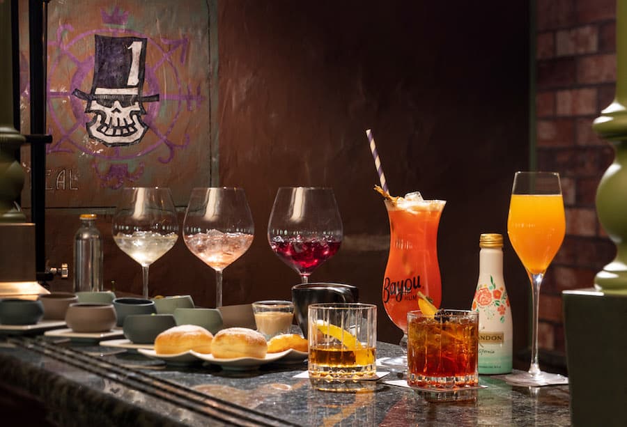 Colorful specialty drinks and whimsical pastries line a counter in a lively, New Orleans-inspired theme park lounge.
