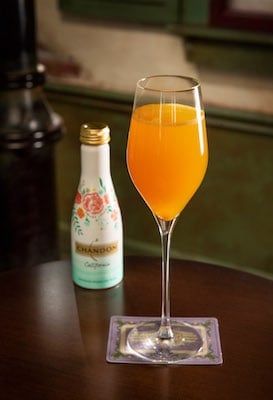 An orange cocktail on a coaster beside a floral bottle atop dark wood, bringing Disney Wonder’s New Orleans flair to your table.