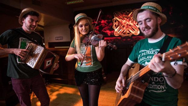 Musicians in festive hats perform guitar, violin, and accordion on Raglan Road’s warmly lit stage for the St. Patrick’s Festival.