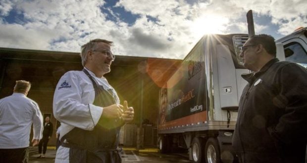 Cast Members chat beside a Disneyland Resorts truck outside a warehouse as surplus food is prepped for the community, sun shining.