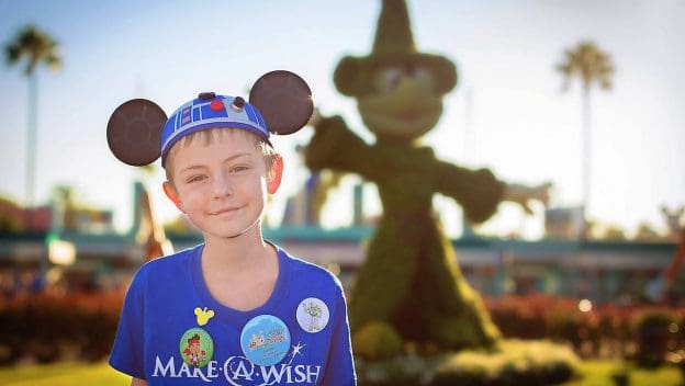 Excited young guest at Disney parks in a Star Wars Mickey hat celebrates World Wish Day near Sorcerer Mickey topiary.