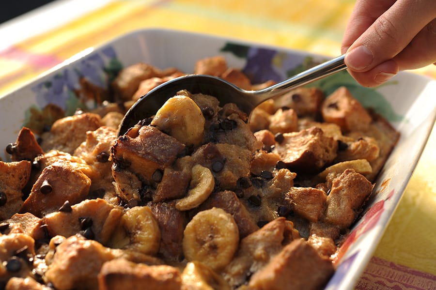 A hand scoops banana bread pudding with chocolate chips from a pan, just like the treat at Disney’s PCH Grill brunch buffet.