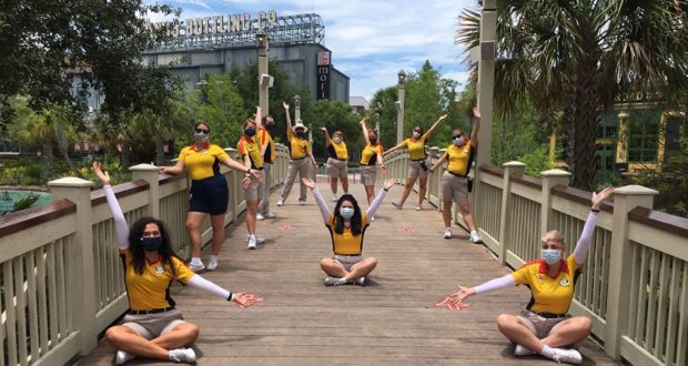 Disney Springs Cast Members in yellow shirts celebrate together on the bridge, masked and socially distanced during reopening.
