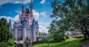 A grand fairytale castle with tall spires and ornate details stands beside a tranquil waterway. Lush green trees and manicured lawns surround the scene under a bright blue sky.