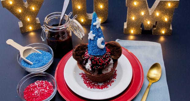 Mickey-themed ice cream sundae topped with a Sorcerer Mickey hat, cookies, sprinkles, and glowing "YUM" sign in the background.