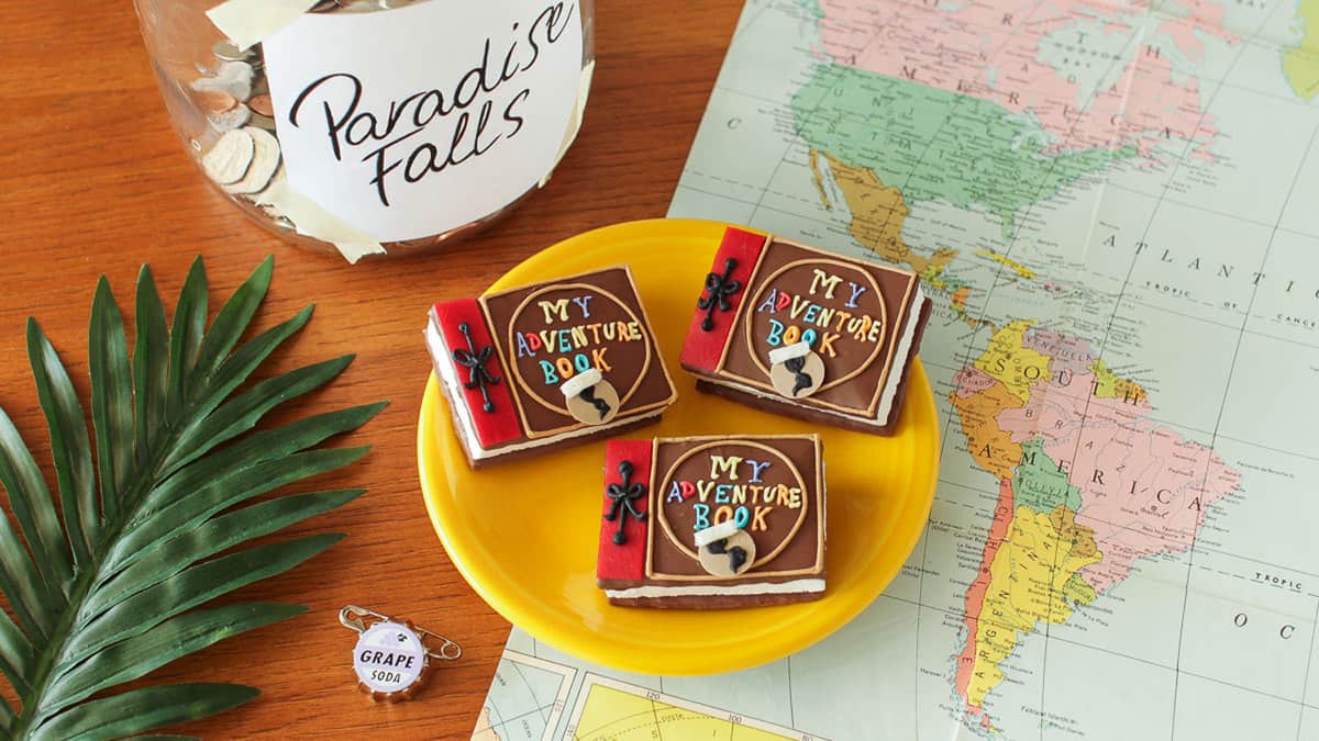 Up-themed "My Adventure Book" cookies on a yellow plate, next to a Paradise Falls jar and map—perfect for Pixar fans on adventure!.
