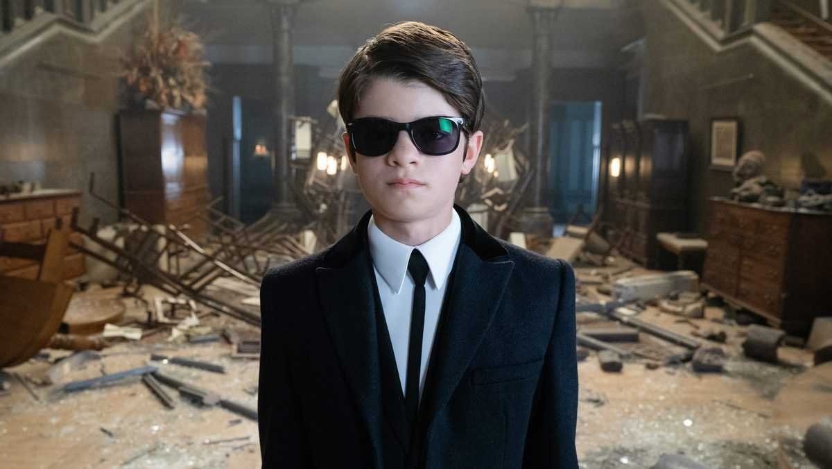 Artemis Fowl, clad in a sleek black suit and shades, stands among wreckage—inviting fans into his thrilling Disney+ world.
